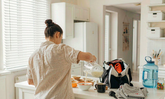 Parent seen from behind pouring breakfast in a sunlit kitchen beside a youth sports bag and gummy jar
