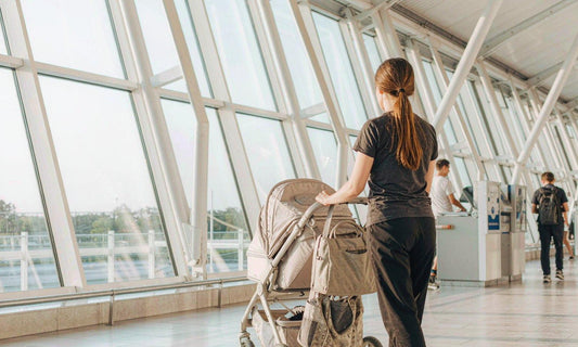 A mother seen from behind pushing a stroller through a sunlit airport terminal with tall glass windows