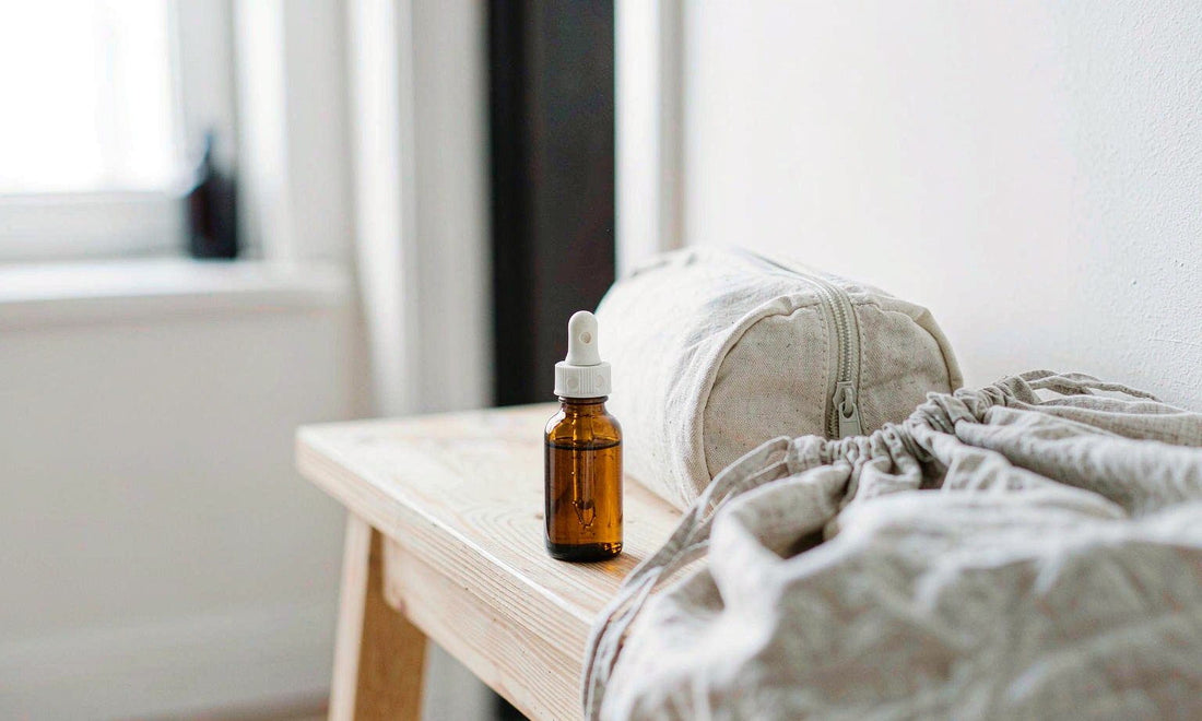 A small amber dropper bottle beside a canvas travel pouch and linen bag on a light wooden bench