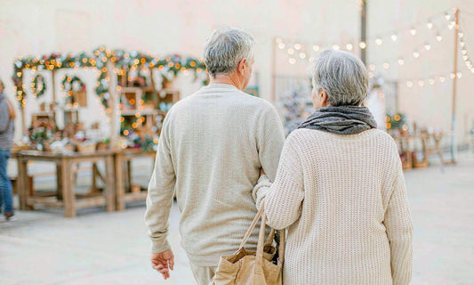 A retired couple seen from behind walking through a bright holiday market with a canvas tote and warm lights