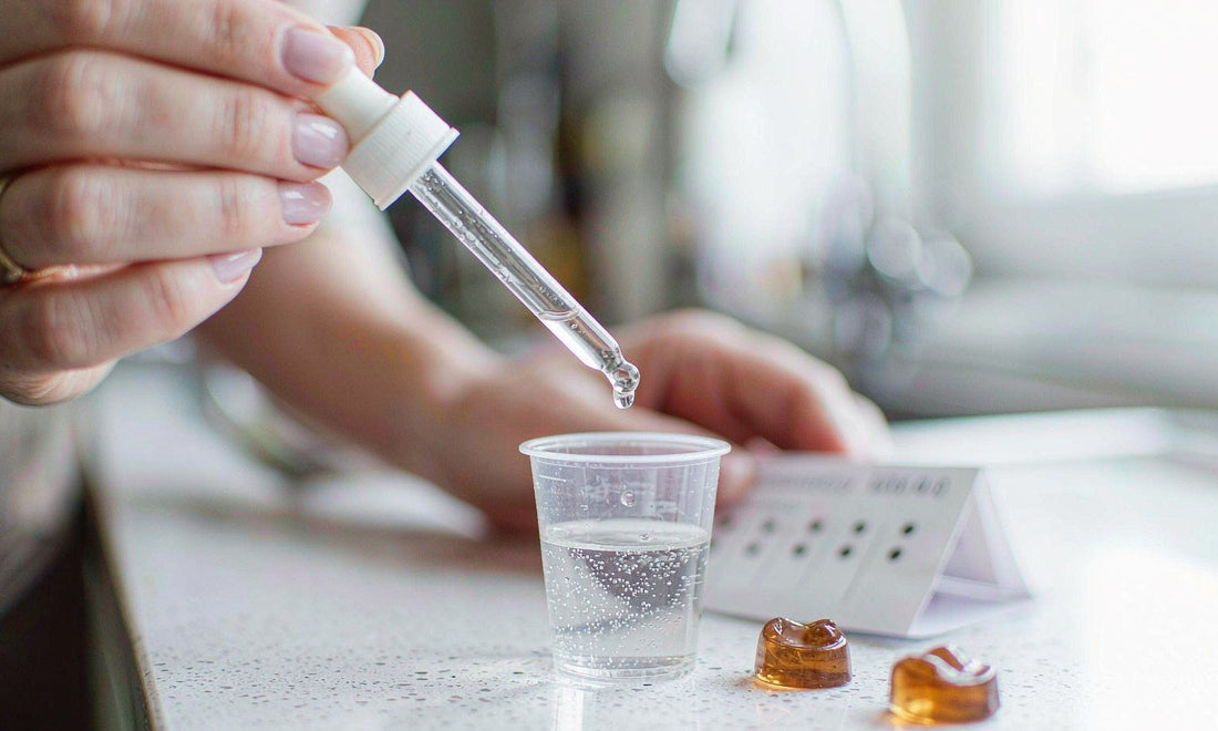A glass dropper filling a small measuring cup beside amber gummies and a folded family list