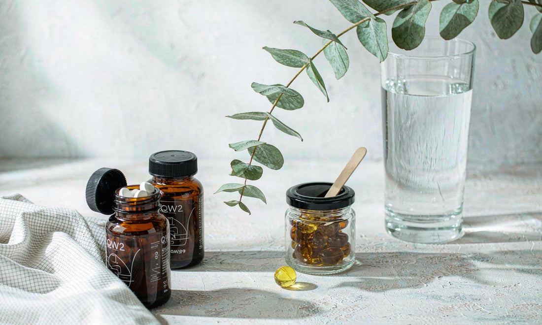 Two amber supplement bottles, a softgel capsule and a gummy jar arranged on pale stone with water