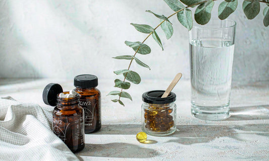 Two amber supplement bottles, a softgel capsule and a gummy jar arranged on pale stone with water