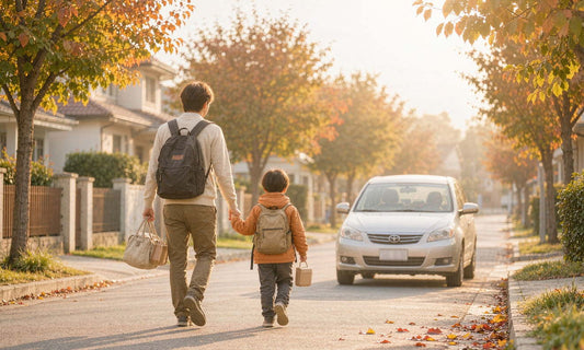 Back view of an adult walking a backpacked child toward a car on a golden autumn school morning