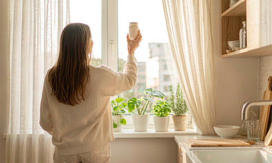 A woman seen from behind in a cream sweater holds a supplement jar up to bright window light in a sunlit kitchen