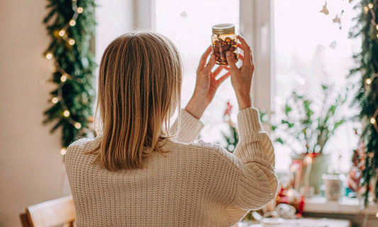 A woman seen from behind in a cream sweater holds a jar of gummies up to window light beside holiday garland