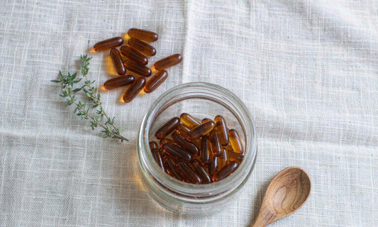 Amber capsules beside an open glass jar, herbs and a wooden spoon on pale linen in soft daylight