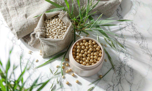 Overhead flat-lay of a supplement jar with golden capsules beside grass blades and a burlap sack of grains on white marble