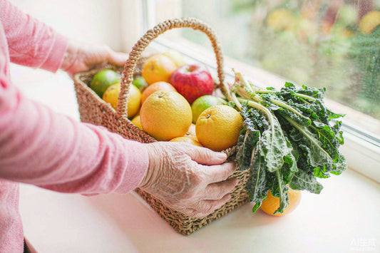 Senior hands placing oranges, apples and leafy greens into a woven market basket on a sunlit kitchen counter