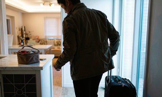 A shift worker seen from behind in silhouette pulling a rolling bag toward a warmly lit kitchen at dawn