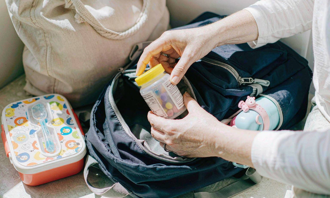 A parent's hands slipping a small gummy jar into a child's backpack beside lunch containers on a bench