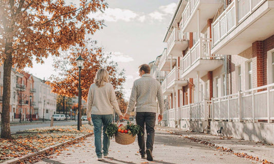 A retired couple seen from behind walking a fallen-leaf sidewalk carrying a market basket of produce