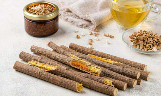 Flat lay of cut licorice root sticks with dried bark, an amber jar and a cup of tea on pale stone