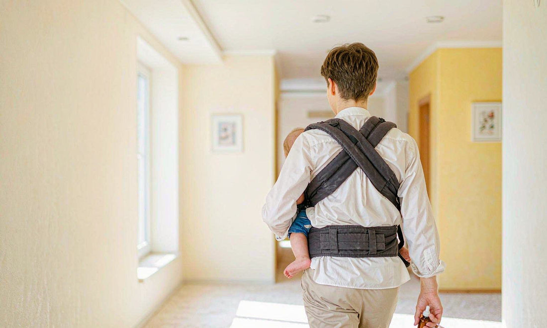 A parent seen from behind carrying a baby in a carrier down a sunlit hallway holding a dropper bottle