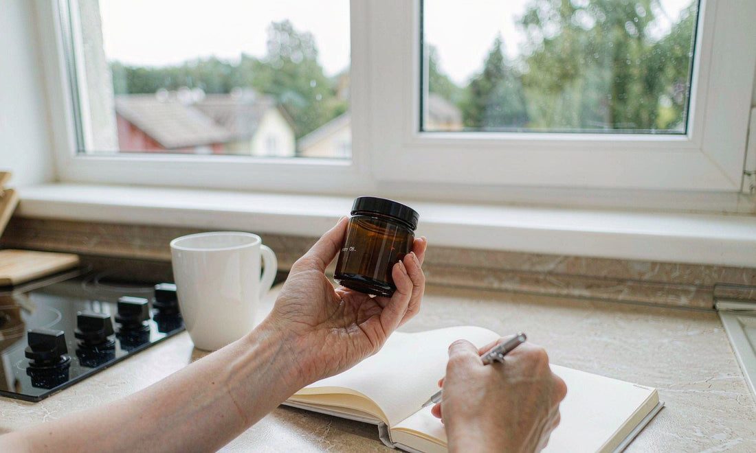 Hands holding a small amber jar beside an open notebook on a bright kitchen counter