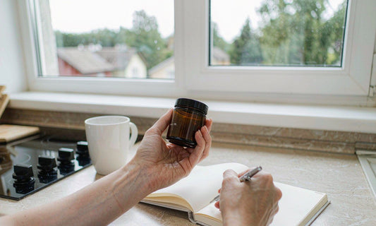 Hands holding a small amber jar beside an open notebook on a bright kitchen counter