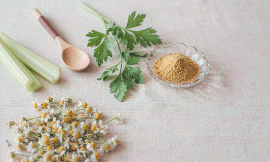 Dried chamomile flowers, fresh parsley and celery with a small dish of golden powder on linen