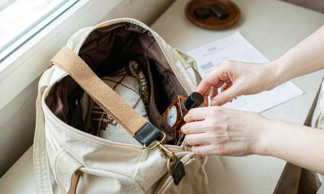 Two hands tucking a small amber drops bottle into a canvas travel bag on an entryway table