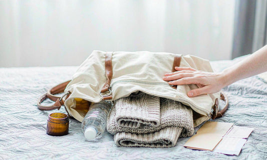 An open canvas travel bag on a pale hotel bed with a water bottle, an amber jar and a folded cardigan