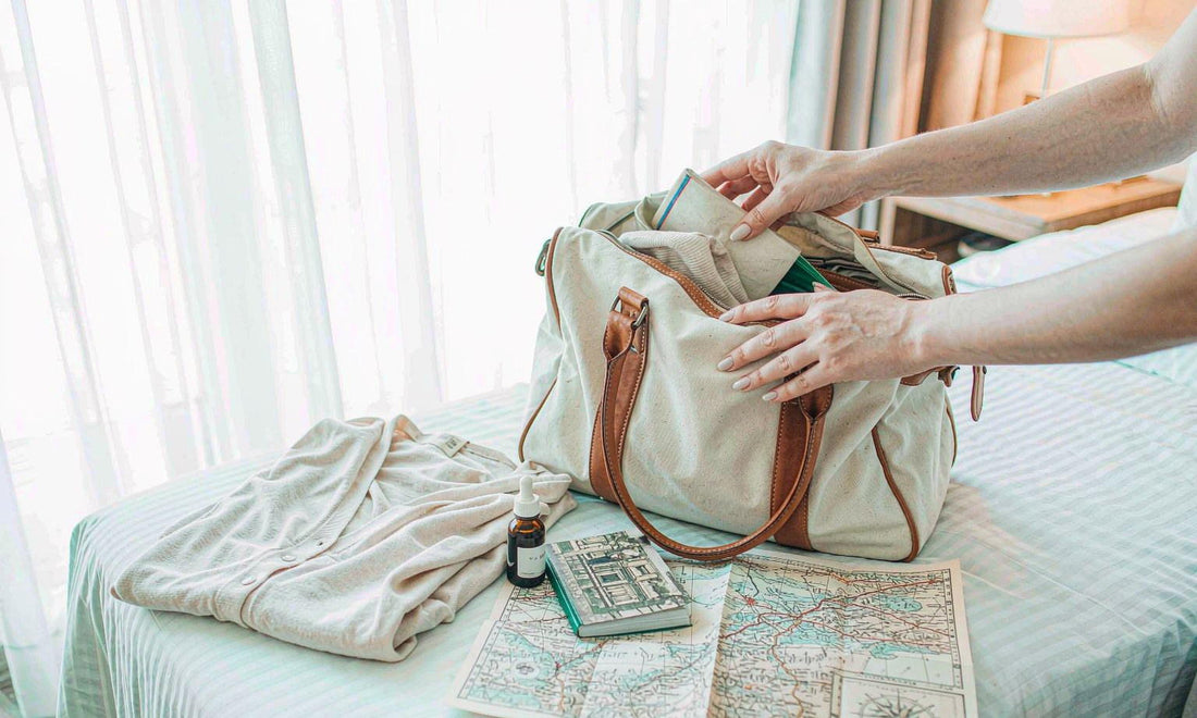 Traveler's hands packing a canvas weekender bag with a dropper bottle and a folded map in bright light