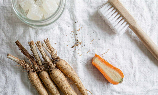 Ingredient flat lay of fresh burdock roots with a wooden brush, carrot and gummy jar on white linen