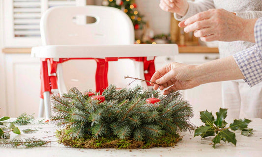 A parent's hands arranging evergreen greenery on a bright kitchen table with a baby high chair behind