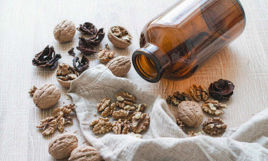 Top-down flat lay of walnuts, dark dried hulls, a plain amber bottle and linen cloth on a pale oak table