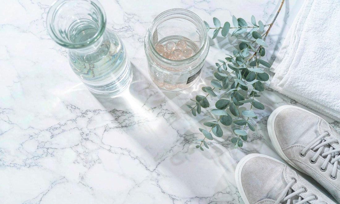 Flat-lay on white marble of a jar of pale gummies beside a water carafe, eucalyptus and walking shoes