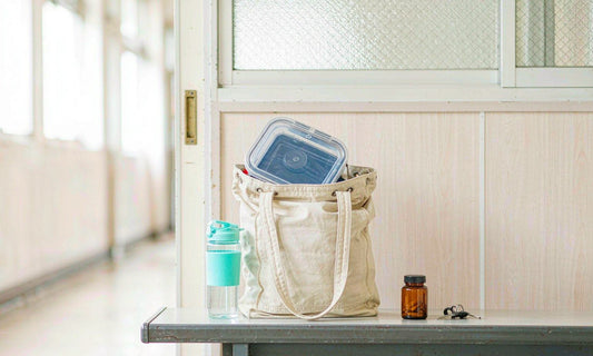 A canvas tote with a lunchbox and water bottle on a hallway bench beside an amber jar and house keys