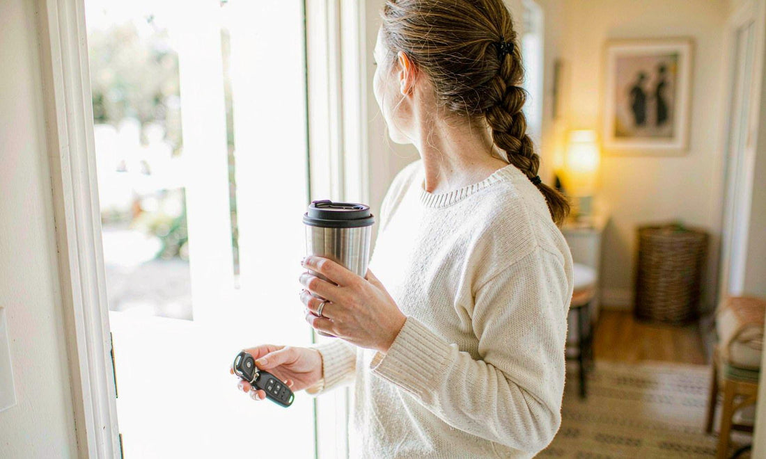 A woman in her thirties holding a travel coffee cup and car keys at a bright open front door