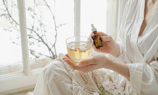 Hands holding a glass of pale herbal infusion and an amber bottle of dried root by a window