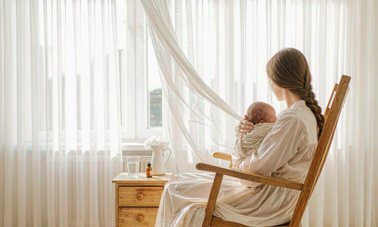 Mother from behind rocking her swaddled newborn by a sunlit window, dropper bottle on the nightstand