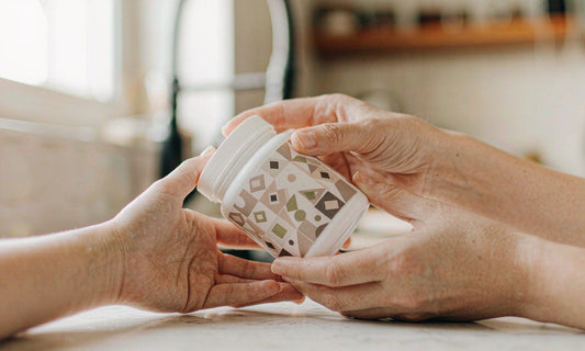 Two hands holding a white supplement jar and reading its label at a sunlit family kitchen counter