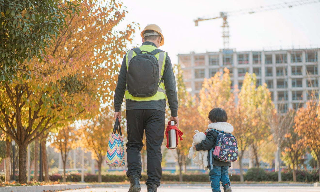 A construction worker seen from behind walking his child to school on a golden autumn morning, backpack on