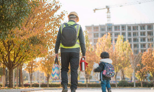 A construction worker seen from behind walking his child to school on a golden autumn morning, backpack on