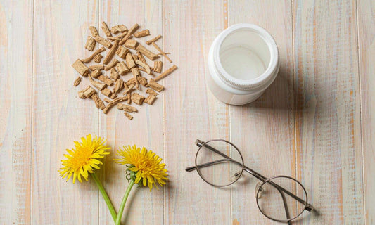 Dried dandelion root, yellow flowers, a white jar and reading glasses on a pale wooden table