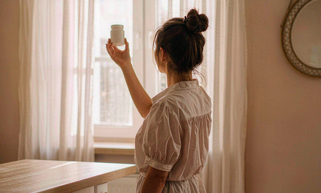 A woman seen from behind holding a white supplement jar up toward a sunny kitchen window