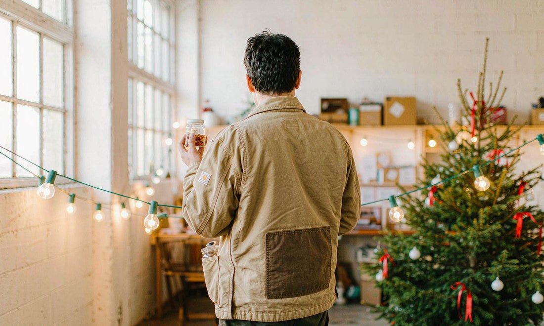 A worker in a canvas jacket seen from behind holding a gummy jar beside holiday lights and an evergreen