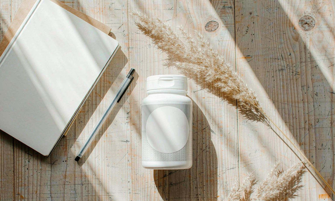 A white supplement bottle with notebook, pen and dried grass on a pale wooden table in morning light