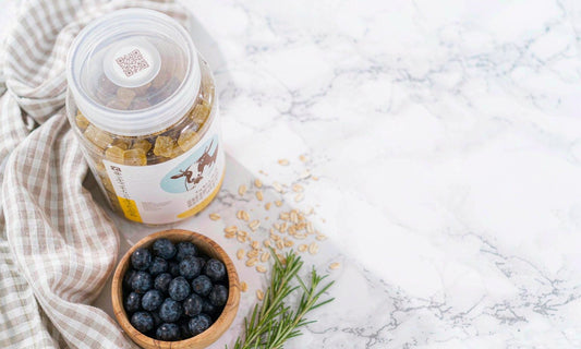 Overhead flat-lay of a gummy jar on pale marble with a wooden bowl of blueberries, oats and rosemary