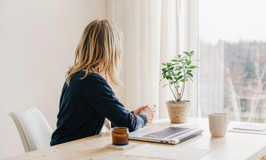 A person seen from behind at a light wood desk with a laptop, an amber jar, a mug and a small plant