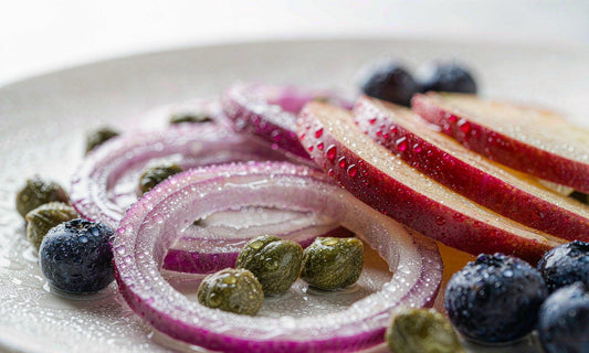 Macro of red onion, apple slices, capers and blueberries on a pale ceramic plate in daylight