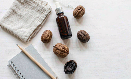 A glass dropper bottle of dark botanical extract with whole walnuts, a linen cloth and a notebook on oak