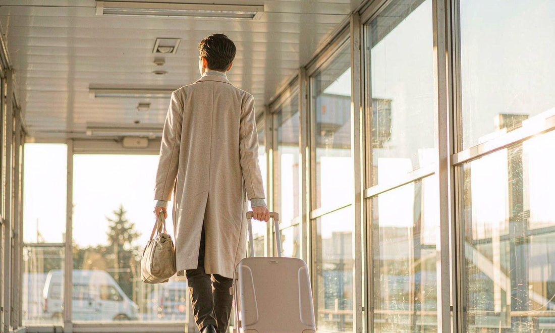 A traveler seen from behind pulling a suitcase through a sunlit airport corridor with a canvas pouch