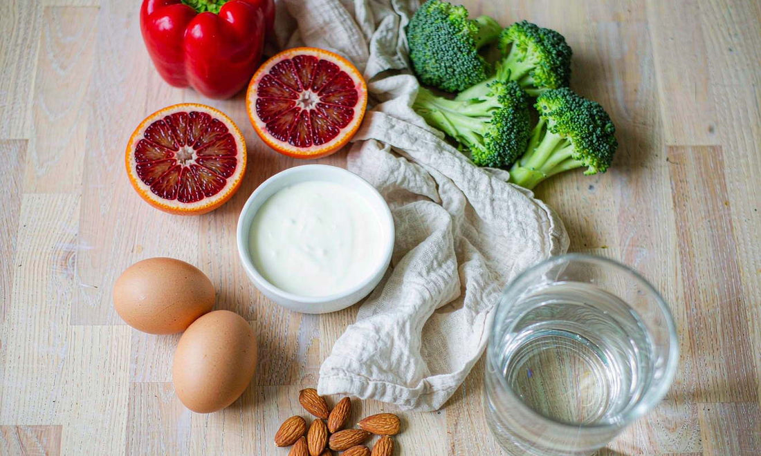 Overhead flat-lay of citrus, peppers, yogurt, eggs and seeds on a pale oak counter