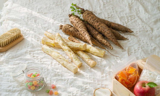Ingredient flat lay of fresh burdock roots with a wooden brush, apple and lunchbox on white linen