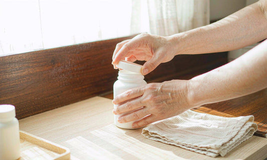 A man's hands screwing the cap onto a white supplement jar on a sunlit pale wood kitchen counter
