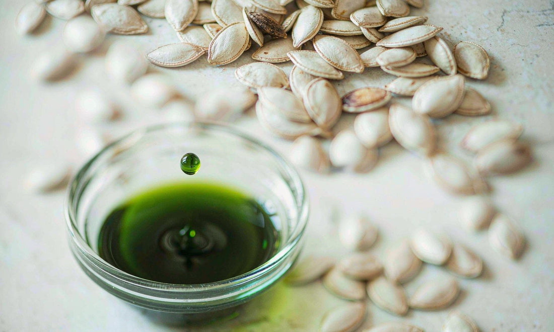 Macro of a dark green pumpkin seed oil drop falling into a small glass bowl beside scattered raw seeds