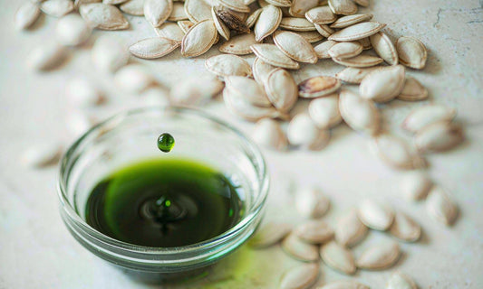 Macro of a dark green pumpkin seed oil drop falling into a small glass bowl beside scattered raw seeds