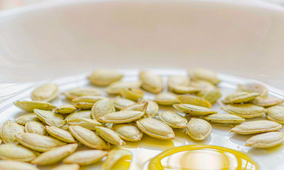 Macro of glossy green pumpkin seeds scattered across white ceramic beside a shallow pool of golden oil